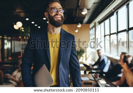 Happy young businessman smiling in a co-working space. Contemplative businessman looking away thoughtfully while holding a laptop. Cheerful young entrepreneur working in a modern workspace.