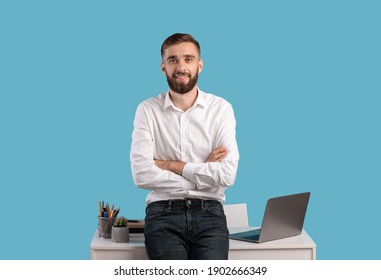 Happy young businessman standing with crossed arms near desk with laptop computer on blue studio background. Millennial entrepreneur smiling and looking to camera at workplace - Powered by Shutterstock