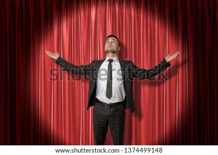 Happy young businessman standing and looking up with arms spread out to the side lit up by spotlight, against red stage curtain. In centre of attention. Gain respect and recognition. Achieve success.