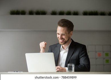 Happy young businessman in suit looking at laptop excited by good news online, lucky successful winner man sitting at office desk raising hand in yes gesture celebrating business success win result - Powered by Shutterstock