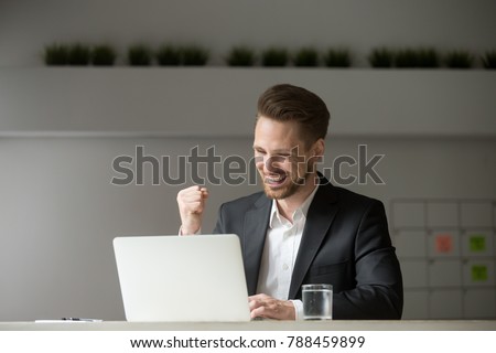 Happy young businessman in suit looking at laptop excited by good news online, lucky successful winner man sitting at office desk raising hand in yes gesture celebrating business success win result
