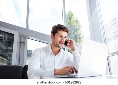 Happy Young Businessman Talking On Cell Phone And Using Laptop In Office