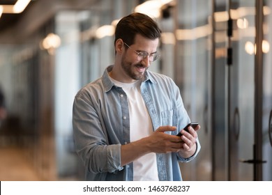 Happy young businessman using smart phone standing in office hallway, smiling male professional holding smartphone working with business apps texting sms enjoy corporate technology on mobile device - Powered by Shutterstock