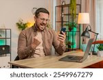 Happy young businessman using smartphone typing browsing saying yes, celebrating success victory, winning lottery jackpot goal achievement good positive news, triumph. Guy at home sitting at table.