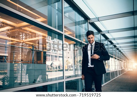 Happy young businessman walking and looking at mobile phone at airport. Handsome business executive texting on smartphone while walking outside airport terminal.