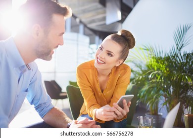 Happy Young Businessman And Woman Sat Working Together In Office With Mobile And Sun Flare