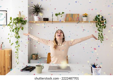 Happy Young Businesswoman Celebrating Success Throwing Confetti Overhead Sitting At Desk In Home Office