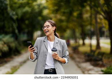 A happy young businesswoman is laughing and holding a cup of coffee while looking at the phone and standing in a park. A woman found something funny on her phone. coffee break - Powered by Shutterstock