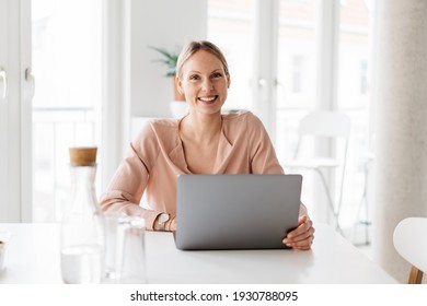 Happy Young Businesswoman With A Lovely Genuine Smile Seated At A Table In A High Key Office Working On A Laptop Computer And Looking At Camera