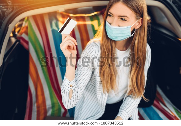 happy young businesswoman in a protective medical\
mask on her face with a credit card in her hand sits in the trunk\
of a car against the background of the sunset, concept of business,\
leisure, travel