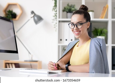 Happy young businesswoman in smart casual looking at you while sitting by desk in front of camera and planning work in office