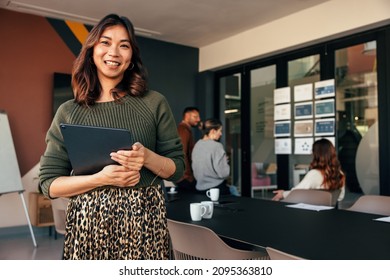 Happy young businesswoman smiling at the camera while holding a digital tablet. Cheerful young businesswoman standing in a boardroom with her colleagues in the background. - Shutterstock ID 2095363810