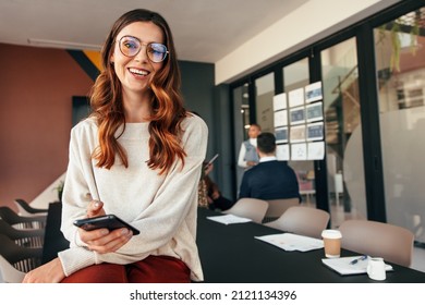 Happy young businesswoman smiling at the camera while holding a smartphone. Cheerful young businesswoman sitting on a boardroom table with her colleagues in the background. - Powered by Shutterstock