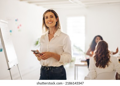 Happy young businesswoman smiling at the camera while holding a smartphone in a meeting room. Cheerful young businesswoman working in an all-female office.