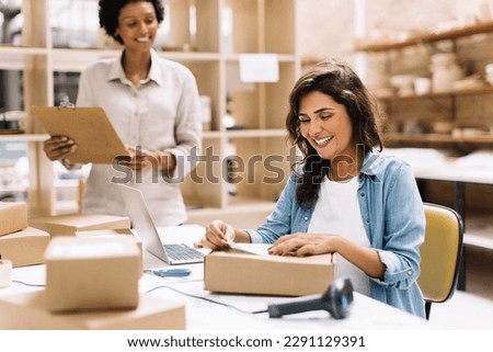 Happy young businesswoman sticking a barcode label on a package box. Online store owners preparing an order for shipping in a warehouse. Female entrepreneurs running an e-commerce small business.