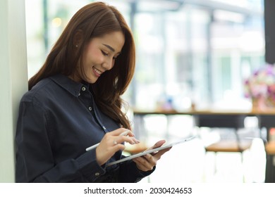 Happy Young Businesswoman Using Portable Tablet Lean On Wall At Office, Chatting With Friends, Scrolling Social Media