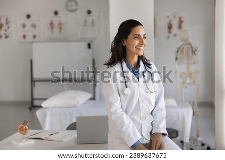 Happy young cardiologist woman posing at workplace, leaning on clinic office table, looking away, smiling, enjoying occupation, thinking on successful doctor career. Casual portrait