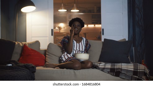 Happy young casual African woman sits on couch at home watching movies on projector with drinks and popcorn slow motion.