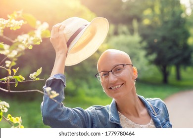 Happy young caucasian bald confident hipster woman take off hat and enjoying life after surviving breast cancer. Portrait of beautiful hairless girl smiling walking city park bright sunny backlit
