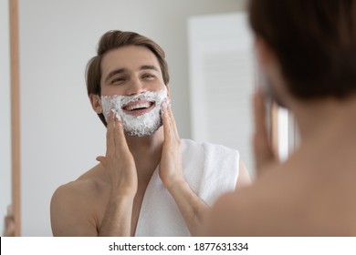 Happy Young Caucasian Bare Man With Towel On Shoulder Applying Shaving Moisturizing Foam On Face, Starting Hygienic Procedure, Smoothing Facial Skin With Mousse, Preparing For Hair Removal Routine.