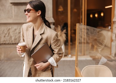 Happy young caucasian brunette woman with laptop and coffee posing outdoors. Girl wears stylish beige jacket, sunglasses. Weekend enjoyment concept - Shutterstock ID 2183162327