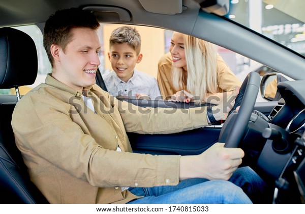 Happy\
young caucasian car owner in dealership,handsome young man sitting\
at front seat of the car, likes design of auto, wife and son\
waiting for his reaction on car, ask about\
feelings