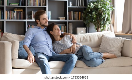 Happy Young Caucasian Couple Relax On Cozy Sofa At Home Hugging Look In Distance Dreaming Together, Dreamy Millennial Man And Woman Rest On Comfortable Couch Embracing, Enjoy Weekend At Home