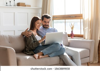 Happy Young Caucasian Couple Rest On Sofa In Living Room Look At Laptop Screen Shopping Online From Home. Millennial Man And Woman Clients Customers Use Modern Computer Gadget On Weekend.