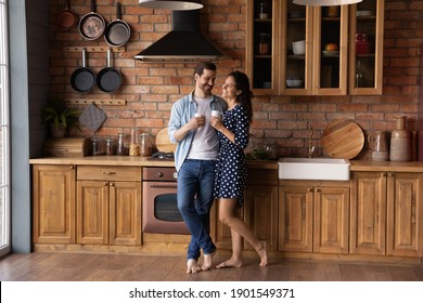 Happy Young Caucasian Couple Stand In Modern Own Kitchen At Home Drink Coffee Or Tea Enjoy Sunny Morning Together. Smiling Millennial Man And Woman Celebrate Relocation Moving To New House.