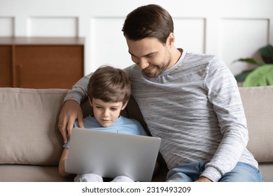 Happy young Caucasian dad sit on sofa with small son use computer gadget learn study online together. Smiling father and little boy child watch funny video on laptop at home. Technology concept. - Powered by Shutterstock