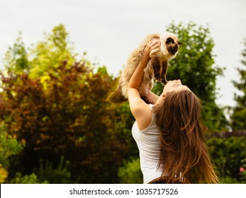 Happy Young Caucasian European Female With White Shirt And Beautiful Brown Hair Holding And Loving A Ragdoll Birma Cat In The Air And Playing On A Garden With Green Trees