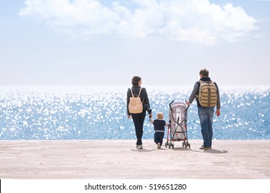 Happy young caucasian family with one year old son and stroller walking along wooden jetty on sunshine, back view. Happy family concept with copy space or text on left area