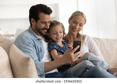 Happy Young Caucasian Family With Small Daughter Relax On Couch Using Smartphone Together. Smiling Mom And Dad Rest On Sofa With Little Girl Child Talk On Video Call On Cellphone Gadget Online.