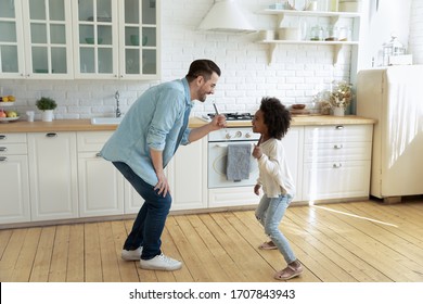 Happy Young Caucasian Father And Little Biracial Daughter Sing In Kitchen Appliances Entertain At Home, Overjoyed Dad And Small African American Girl Child Have Fun Play On Weekend Together