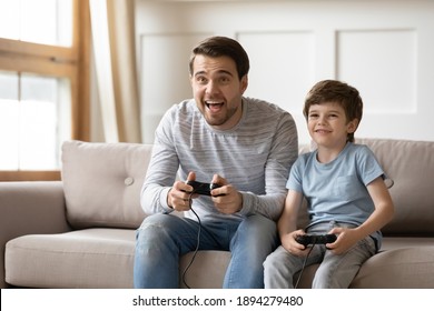 Happy Young Caucasian Father And Little 6s Son Sit On Couch At Home Have Fun Playing Computer Video Game Together. Overjoyed Dad And Small Boy Child Enjoy Family Weekend Engaged In Funny Activity.