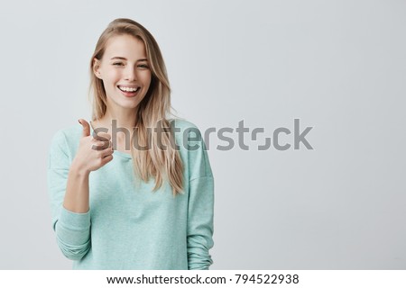 Happy young caucasian female wearing blue long sleeved shirt making thumb up sign and smiling cheerfully, showing her support and respect to someone. Body language. I like that. Good job.