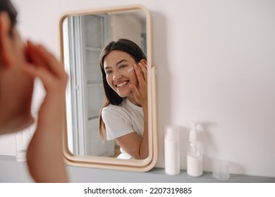 Happy young caucasian girl applying skincare product to face in front of mirror at home. Brunette hair woman using facial cream indoor. Concept beauty - Shutterstock ID 2207319885