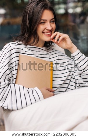 Happy young caucasian girl with notebook smiling looking away sitting outdoors. Brunette wearing sweatshirt spends her leisure time alone. Sincere emotions lifestyle concept