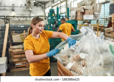 Happy young caucasian girl putting disposable cups into bag while working on waste dumping station. Garbage sorting and recycling concept