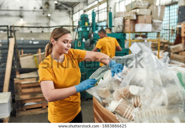 Happy young caucasian girl putting disposable\
cups into bag while working on waste dumping station. Garbage\
sorting and recycling\
concept