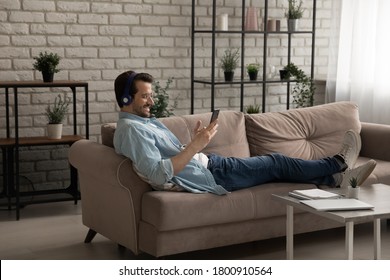 Happy Young Caucasian Man In Earphones Sit Relax On Couch In Living Room Browse Internet On Modern Cellphone Gadget, Smiling Millennial Male In Headphones Listen Music On Smartphone At Home