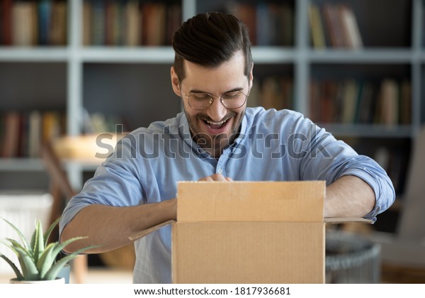 Happy young Caucasian man feel excited unbox\
unpack cardboard box shopping online from home. Smiling millennial\
male buyer triumph satisfied with good product quality. Delivery\
service concept.