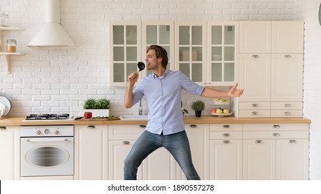 Happy Young Caucasian Man Have Fun Sing In Appliances Dance In Cozy Modern Renovated Kitchen. Smiling Millennial Male Renter Enjoy Cooking At Home, Celebrate Relocation To New Own House Or Flat.