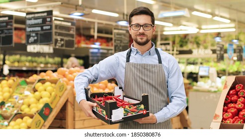 Happy young Caucasian man seller in good mood standing in supermarket with box with red hot pepper and looking at camera. Female in apron working at grocery. Portrait concept. Retail business - Powered by Shutterstock