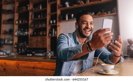 Happy Young Caucasian Man Sitting At Cafe Making Video Call From His Mobile Phone. Caucasian Male At Coffee Shop Having A Videochat On Smart Phone.