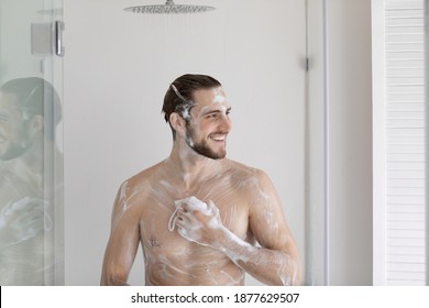 Happy young caucasian man standing under pouring shower, cleaning body with foamy gel, exfoliating skin using washcloth, feeling energetic enjoying morning hygienic cleansing routine in bathroom.