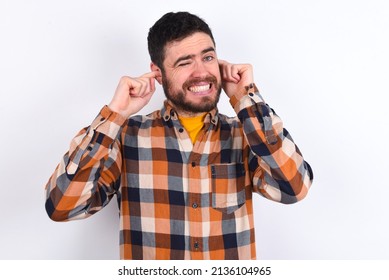 Happy Young Caucasian Man Wearing Plaid Shirt Over White Background Ignores Loud Music And Plugs Ears With Fingers Asks To Turn Off Sound