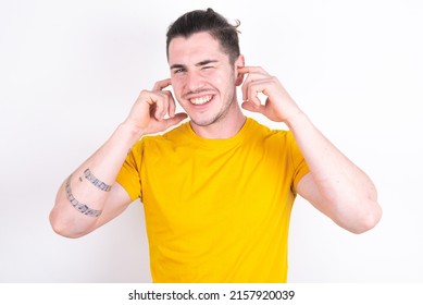 Happy Young Caucasian Man Wearing Yellow T-shirt Over White Background Ignores Loud Music And Plugs Ears With Fingers Asks To Turn Off Sound