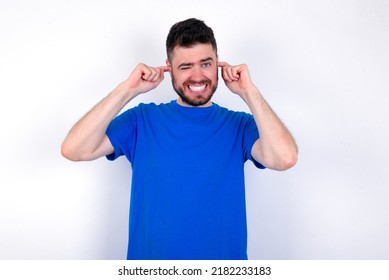 Happy Young Caucasian Man Wearing Blue T-shirt Over White Background Ignores Loud Music And Plugs Ears With Fingers Asks To Turn Off Sound