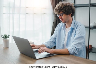 Happy Young Caucasian Man Wearing Glasses And Smiling Working Typing On His Laptop To Get All His Business Done Early In The Morning With His Cup Of Coffee. Young White Man Studying Online At Home.